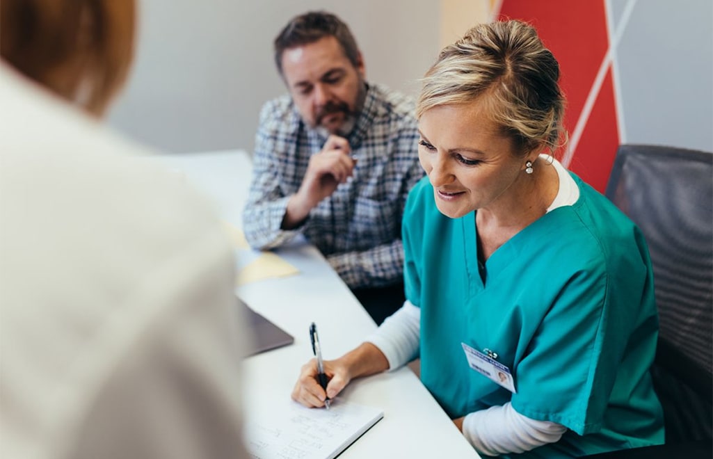 A female health care worker is seated writing on a piece of paper beside a man